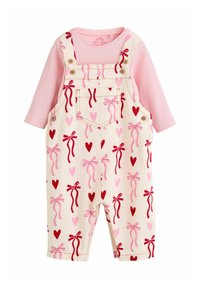 Roza majica z dolgimi rokavi pod krem kombinirano obleko z vzorci rdečih in roza pentelj ter srčkov. Material je videti mehak; ima nastavljive naramnice in žepke.