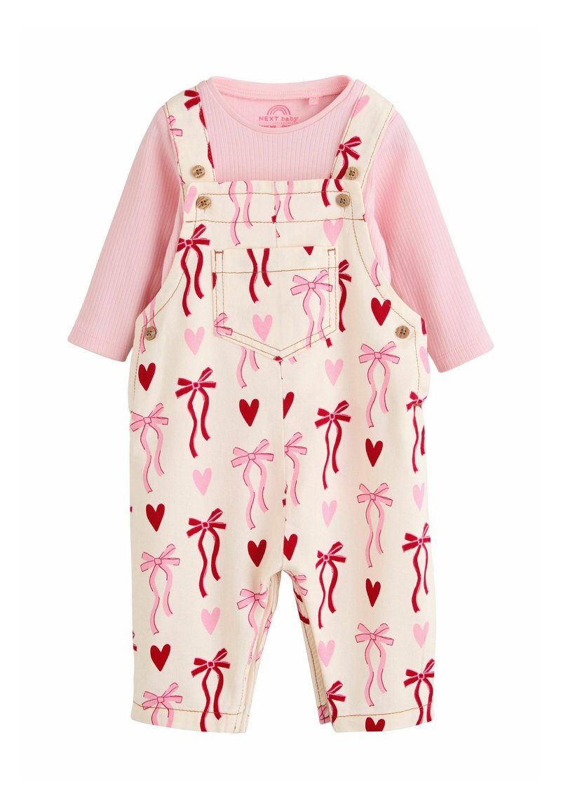 Roza majica z dolgimi rokavi pod krem kombinirano obleko z vzorci rdečih in roza pentelj ter srčkov. Material je videti mehak; ima nastavljive naramnice in žepke.