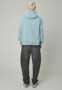 Personne avec les cheveux blonds courts portant un sweat à capuche bleu clair, un jean gris foncé ample et des baskets, se tenant de dos contre un fond uni.