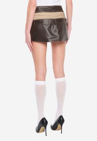 Mini-jupe en similicuir noir avec une ceinture beige, présentant des détails de couture, associée à des chaussettes blanches hautes jusqu'aux genoux et des escarpins noirs.