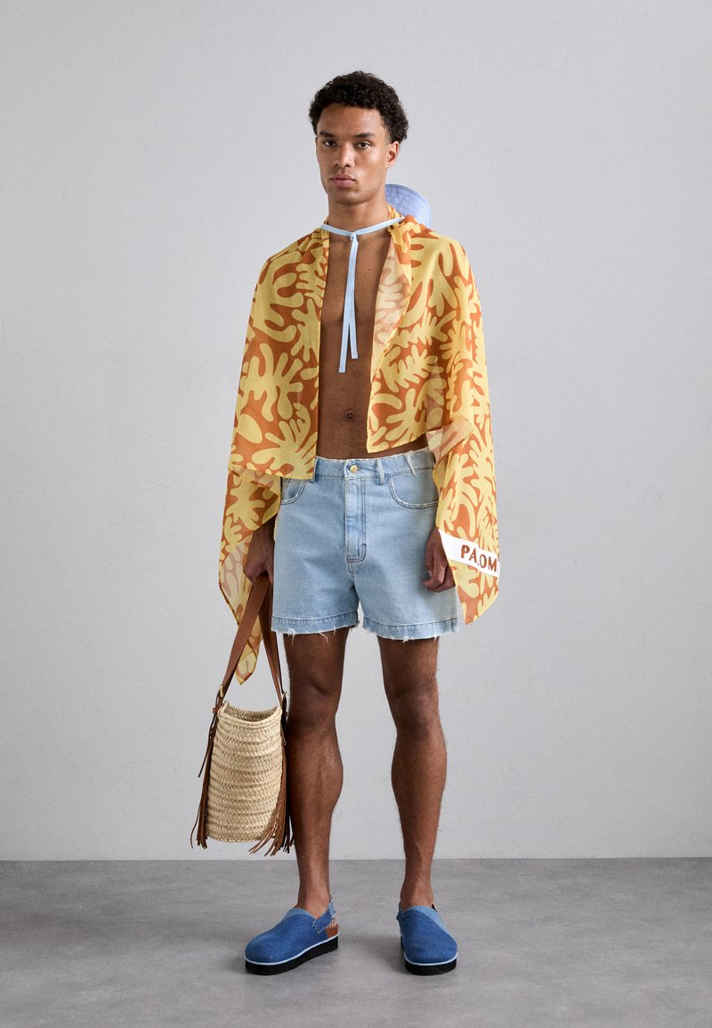 Lightweight yellow and orange patterned cape, denim shorts, blue clogs, and a woven bag with a fringe detail.