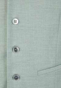 Hellgrüner Stoff mit einer strukturierten Webart, versehen mit vier metallischen Knöpfen und einem kleinen Taschen-Detail auf der linken Seite.