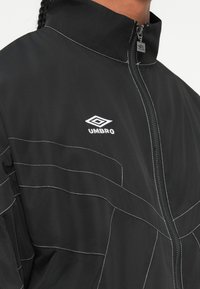 Kadrova snimka crne jakne na zatvaranje s patentnim zatvaračem s bijelim kontrastnim šavovima i bijelim Umbro logotipom na prsima, koju nosi osoba s pletenicama.