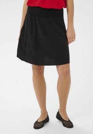 Žena v černé sukni s elastickým pasem, červeném topu a černých pletených balerínkách stojí na bílém pozadí.