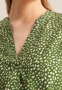 Zelený textilní top s výstřihem do V, s texturovaným vzorem bílých a světlezelených teček. Detailní pohled na výstřih a potisk.