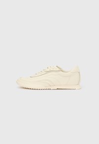 Madala profiiliga kreemivärvi toss, millel on laineline õmblus, paelaga kinnitamine ja sooneline tallaosa, nähtav küljelt ühtlasel taustal.