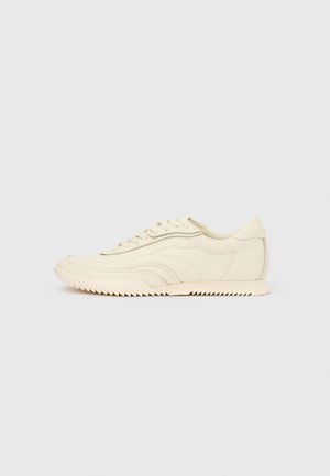 Lavprofileret sneaker i cremefarve med bølget syning, snørebånd og en rilleret sål, vist fra siden på en ensfarvet baggrund.