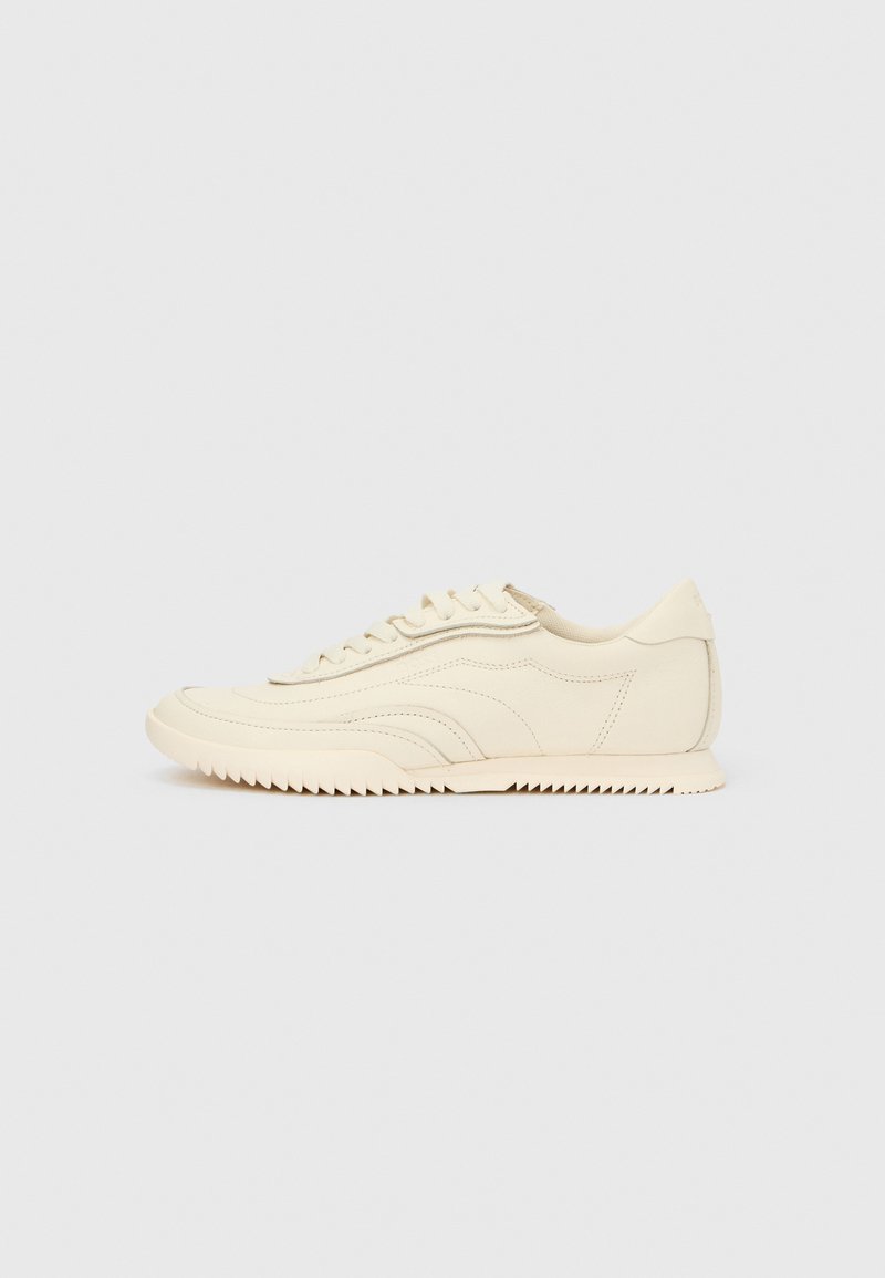 Madala profiiliga kreemivärvi toss, millel on laineline õmblus, paelaga kinnitamine ja sooneline tallaosa, nähtav küljelt ühtlasel taustal.