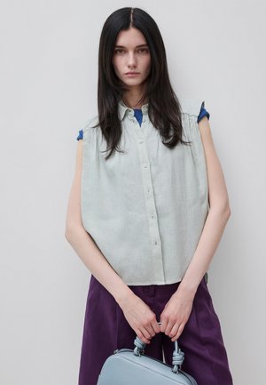 Vrouw met lang donker haar die een licht mouwloos knoopshirt en paarse broek draagt, houdt een lichtblauwe handtas vast voor een grijze achtergrond.