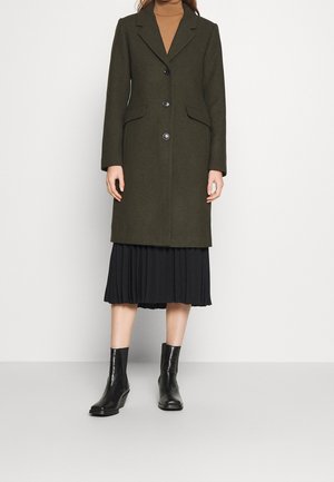 Femme portant un manteau boutonné vert foncé sur une jupe plissée noire, un haut marron à col montant et des bottines noires sur un fond uni.