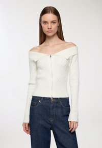 Biała, sweter z dekoltem off-the-shoulder w sploty warkoczowe z prążkowanymi detalami i zamkiem błyskawicznym z przodu, w połączeniu z ciemnoniebieskimi dżinsami.