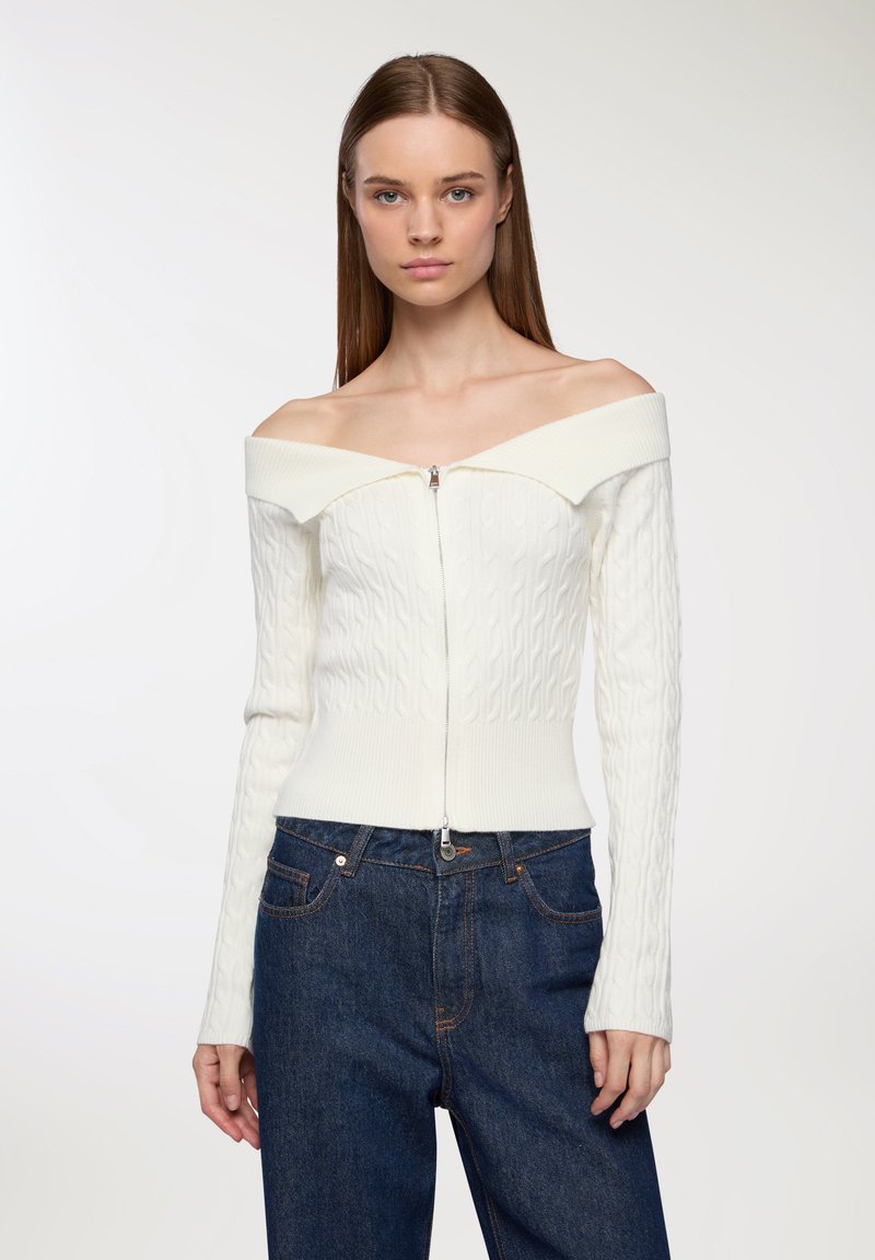 Biała, sweter z dekoltem off-the-shoulder w sploty warkoczowe z prążkowanymi detalami i zamkiem błyskawicznym z przodu, w połączeniu z ciemnoniebieskimi dżinsami.