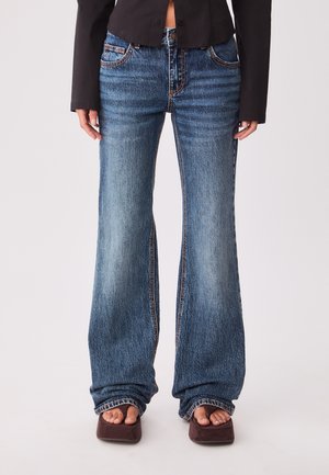 Kvinde iført højtaljede, falmede blå flared jeans kombineret med brune åben-tåede sko med firkantet hæl og en kortsort jakke.