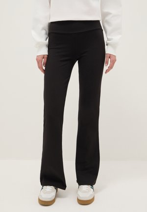 Leggings neri a zampa realizzati in tessuto elasticizzato, con vita alta e texture liscia. Indossati con sneakers bianche.