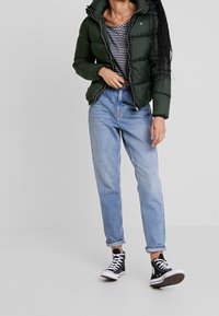 Person som bär en mörkgrön puffjacka, en svart-vitt randig topp, ljusblå uppvikta jeans och svarta hög-topp sneakers som står mot en enfärgad bakgrund.