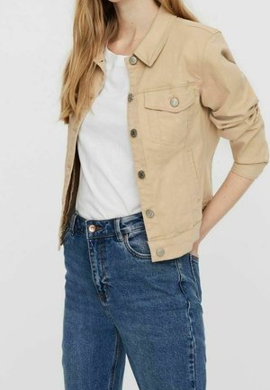Vrouw draagt een beige jasje met knopen over een wit t-shirt en blauwe spijkerbroek, met handen op de heupen tegen een effen achtergrond.
