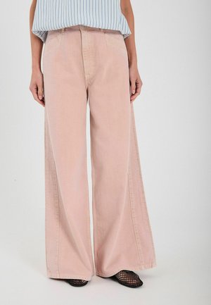 Jasnoróżowe dżinsy z szerokimi nogawkami, wykonane z bawełny, z frontowymi kieszeniami i widocznym przeszyciem, w połączeniu z niebieską bluzką w paseczki. Widoczne czarne buty z siateczki.