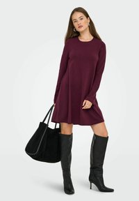 Femme portant une robe bordeaux à manches longues, tenant un grand sac fourre-tout noir, accompagnée de bottes à talons hauts noires jusqu'aux genoux et de boucles d'oreilles en argent.