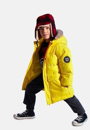Żółta puchowa kurtka z kapturem wyłożonym futrem, czarny pikowany wzór na rękawach, zestawiona z ciemnymi dżinsami i czarnymi trampkami z białymi podeszwami.