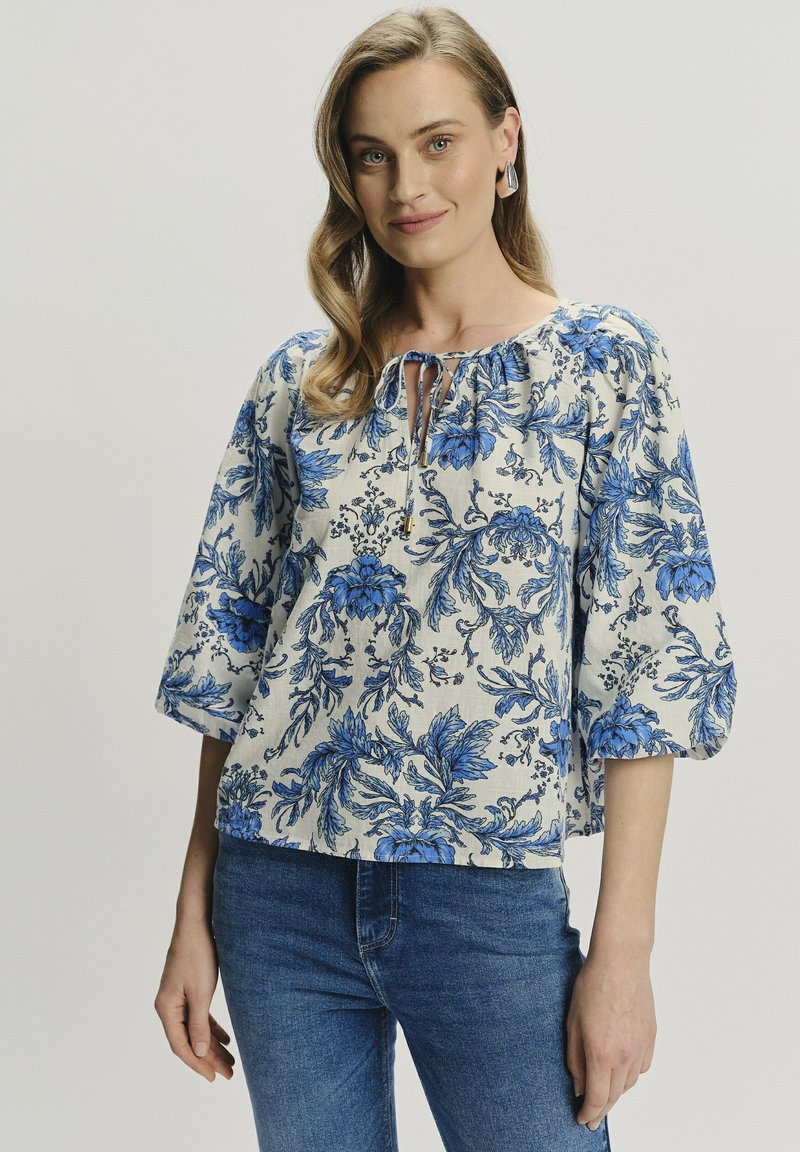 Blusa floreale in crema con motivi blu, caratterizzata da un collo arruffato e maniche a 3/4; abbinata a jeans in denim.