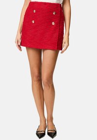 Red textured mini skirt with double-breasted design, featuring gold buttons and a straight hem. Worn with black pointed-toe shoes.