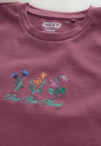T-shirt violet à coupe décontractée avec trois fleurs brodées en point de croix et texte en cursive bleu sarcelle « Flash Spast Atwuit » sous les motifs.