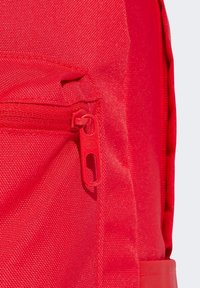 Mochila roja con un tejido texturizado, cremallera prominente con un tirador alargado y un bolsillo lateral. Diseño simple y minimalista.