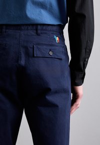 Pantaloni din bumbac de culoare bleumarin, cu un buzunar pe spate prevăzut cu o închidere cu nasture și un logo cu un cal brodat multicolor. Textură netedă, croială ajustată.