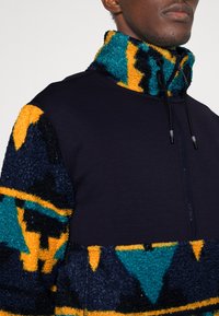 Pulls en polaire avec un tissu bleu marine texturé et des motifs abstraits vibrants en orange, teal et noir. Col montant avec cordons de réglage.