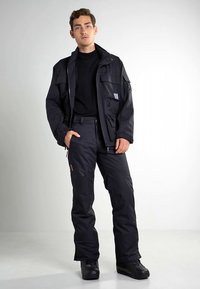 Casaco impermeável preto com múltiplos bolsos, combinado com calças pretas a condizer. Textura suave, fechos funcionais e calçado resistente.