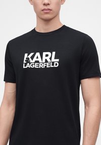 Άνδρας που φοράει μαύρο T-shirt με λευκό λογότυπο "KARL LAGERFELD" και μικρό σιλουέτα γραφικό στο στήθος, εμφανίζεται από το λαιμό μέχρι τη μέση.
