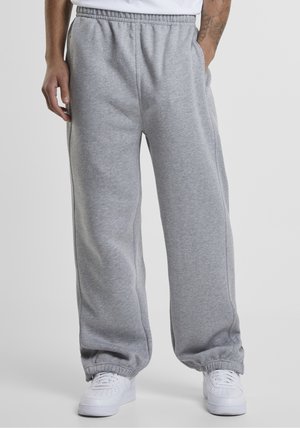 Mężczyzna w luźnych szarych spodniach dresowych z elastycznymi mankietami i białych tenisówkach stojący na jednolitym tle.