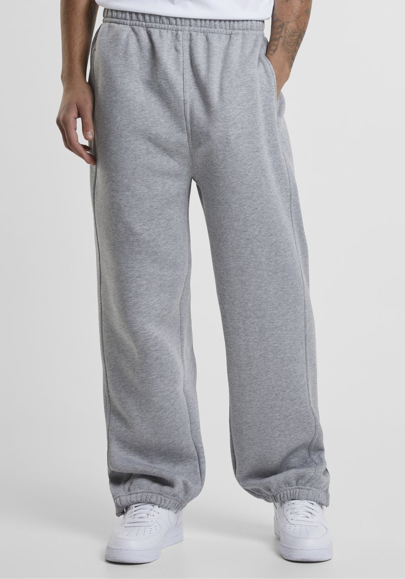 Mees, kes kannab lahtisi halli dressipükse elastsete varrukatega ja valgeid tossusid, seisab lihtsa tausta ees.