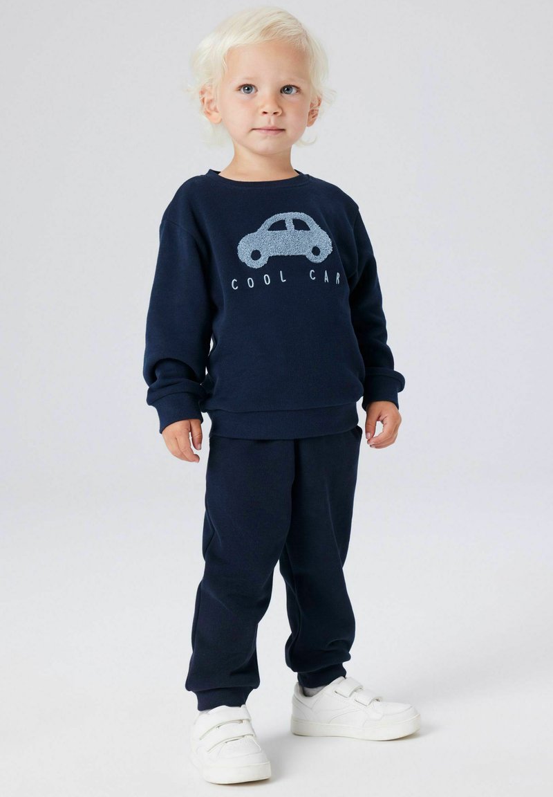 Marineblå genser og bukse sett. Genseren har et teksturert bildesign med teksten "COOL CAR". Barnet har på seg hvite sko med borrelås.