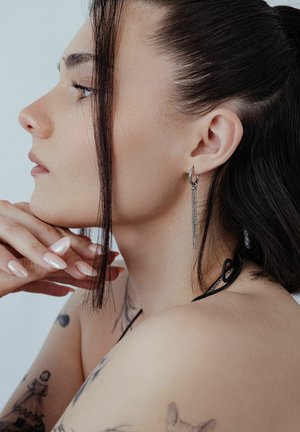 Junge Frau mit dunklem, zurückgebundenem Haar, die ihr Kinn auf die Hand stützt, zeigt Ohrringe, helle Tätowierungen an Schulter und Arm und lange Nägel.