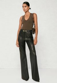 Kobieta w bezrękawnikowej brązowej bluzce, wysokich czarnych skórzanych spodniaczkach z szerokimi nogawkami i paskiem, nosząca brązową torbę na ramię, stojąca przy jasnej ścianie.