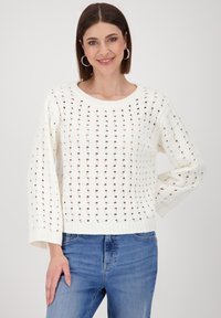White knitted sweater featuring a loose, open weave pattern, wide sleeves, and a ribbed hem. Paired with blue denim jeans.