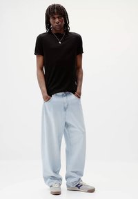 Tricou negru cu mânecă scurtă asociat cu blugi evazați de culoare albastru deschis și adidași gri cu accente purpurii. Modelul stă cu încredere, cu mâinile în buzunare.