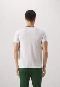 Bílý sportovní triko s kulatým zeleným výstřihem, krátkými rukávy a hladkým povrchem. Oblékané se zelenými sportovními kalhotami s bočními detaily.