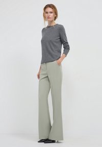 Femme portant un pull gris à manches longues et un pantalon large gris clair, se tenant avec une main dans la poche devant un fond blanc uni.