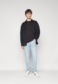 Chaqueta ligera negra con dos bolsillos en el pecho, usada sobre una camisa negra; combinada con jeans de mezclilla azul claro y zapatillas blancas con detalles en negro.