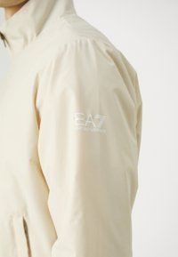 Giacca leggera beige realizzata in materiale liscio, dotata di un colletto alto e del logo EA7 Emporio Armani in bianco sulla manica sinistra.