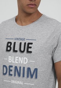 Camiseta de algodón gris con cuello redondo, que presenta un texto en negrita: "DENIM ORIGINAL MEZCLA AZUL VINTAGE" en negro, azul y blanco. Textura suave.