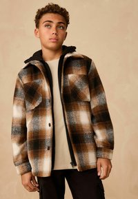 Brown and black plaid flannel jacket with a hood, featuring two chest pockets, textured fabric, and a zip front over a beige shirt.