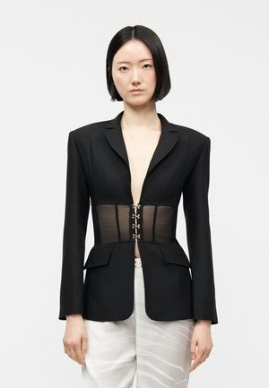 Donna con capelli neri e lisci indossa un blazer nero con un corpetto trasparente in stile corsetto e pantaloni bianchi con un motivo sottile, in piedi davanti a uno sfondo semplice.