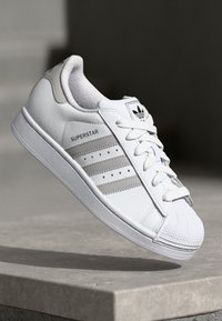 White leather sneaker with three grey stripes, round toe, rubber sole, and textured toe cap. Features branding on the tongue and side.
