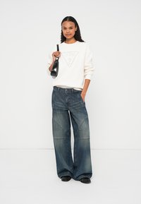 Tröja i ljus krämfärg med präglad logotyp; lediga mörkblå denimjeans med vida ben och tydliga söm- och stygn detaljer; svart väska.