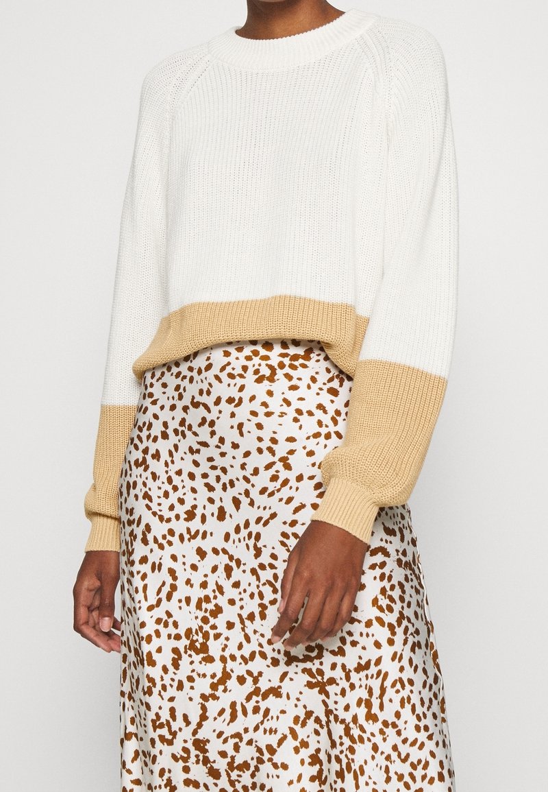 Personne portant un pull blanc avec des poignets et un ourlet beige, associé à une jupe blanche avec un motif de taches brunes irrégulières.
