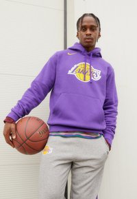 Felpa viola con logo dei Los Angeles Lakers, tasca a marsupio e dettagli gialli; pantaloni della tuta grigi con logo dei Lakers; tiene un pallone da basket.
