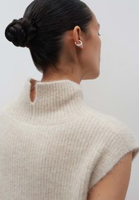 Femme aux cheveux foncés, avec un chignon tressé, portant un pull sans manches en crème à côtes et des boucles d'oreilles en forme de croissant argenté, sur fond blanc.